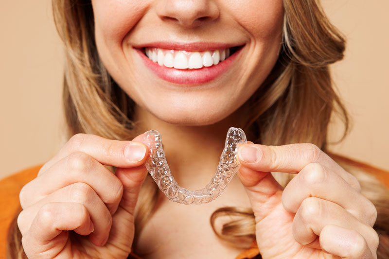a woman smiling and holding up an invisalign clear aligner that highlights her straight teeth